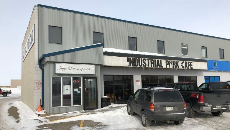 A low-rise building clad in alumninium siding has a large sign that reads 'Industrial Park Cafe.' Some of the letters are formed by the images of tools.