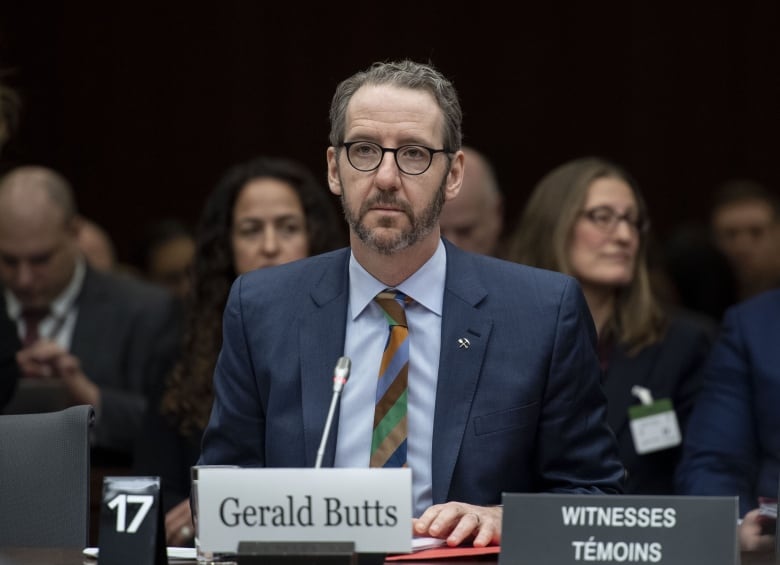 A man with grey hair and glasses sits at a desk with a microphone.