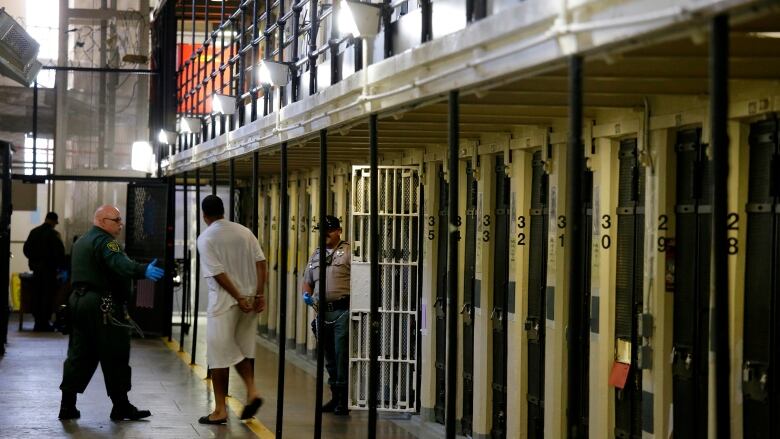 A uniformed prison guard wearing rubber gloves gestures at a prisoner with his hands cuffed behind his back to follow him. A row of cells runs along the right wall and another guard stands beside an open cell door. 