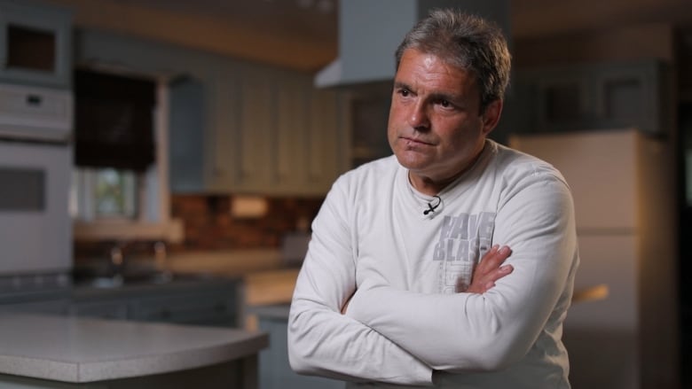 A man with grey hair, wearing a white shirt, is sitting with his arms crossed.