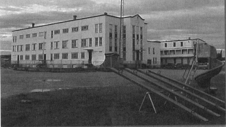 Black and white photo of a large imposing institutional building with swingsets in the foreground. 