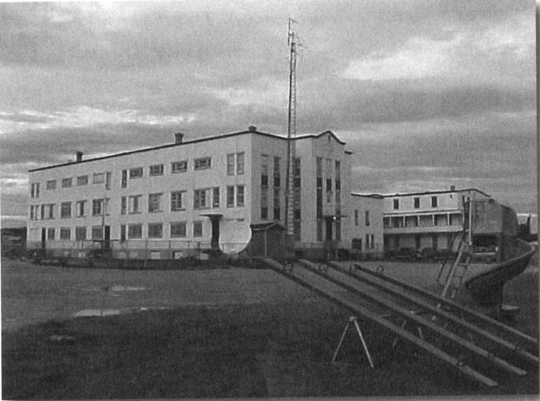 Black and white photo of a large imposing institutional building with swingsets in the foreground. 