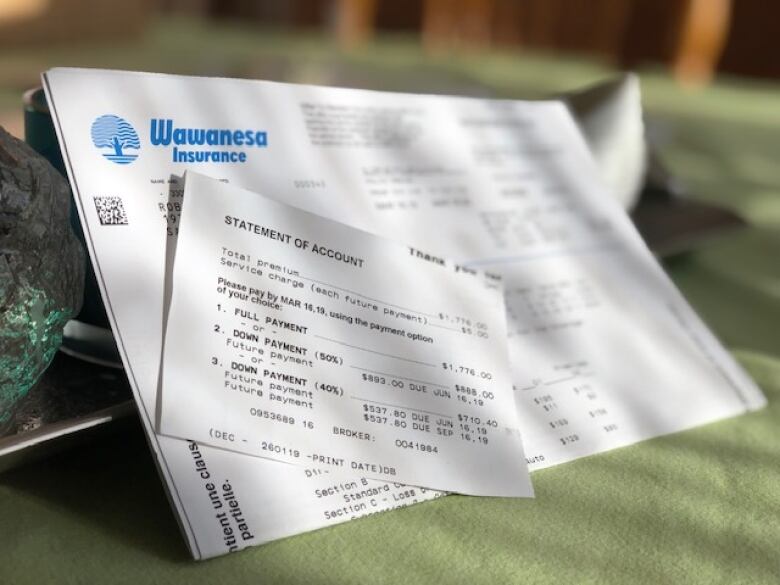 An invoice from Wawanesa Insurance is displayed on a cloth background. A small paper saying Statement of Account obscures personal details of the client. 