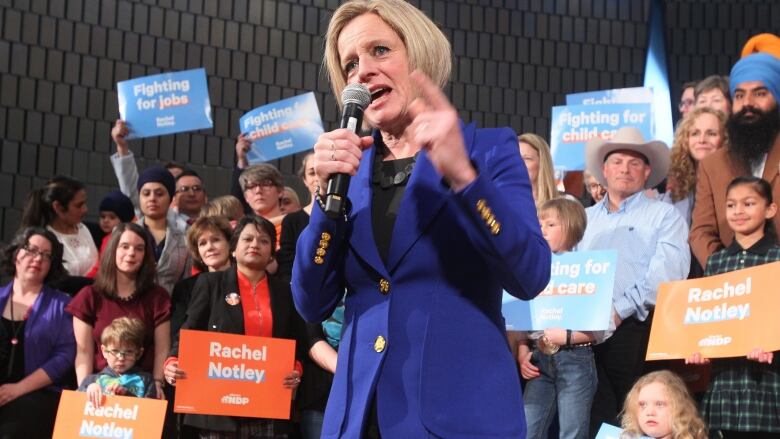 A politician speaks into a microphone, with sign-waving people behind her. 