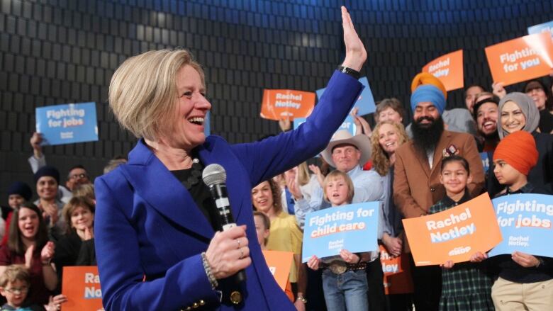 Rachel Notley in a blue suit and holding a microphone waves to a crowd of supporters.