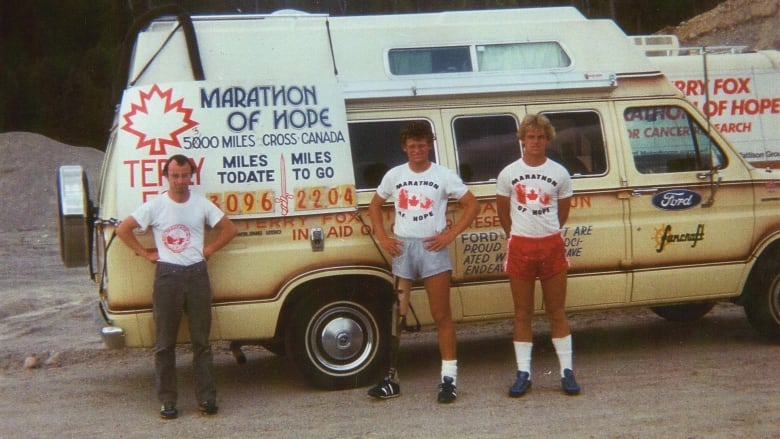 Three men stand in front of a beige van. 