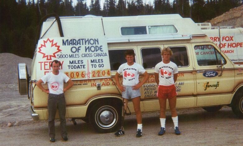 Three men stand in front of a beige van. 