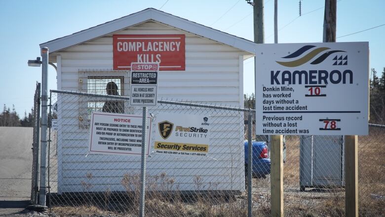 A man is seen inside a guard house behind a chain link fence with a sign saying complacency kills and another saying the mine worked 10 days without a lost time incident.