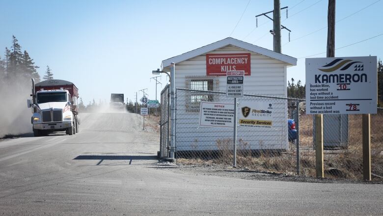 Two large trucks pass on a dusty road leading past the gate and guardhouse at the entrance to a mine.