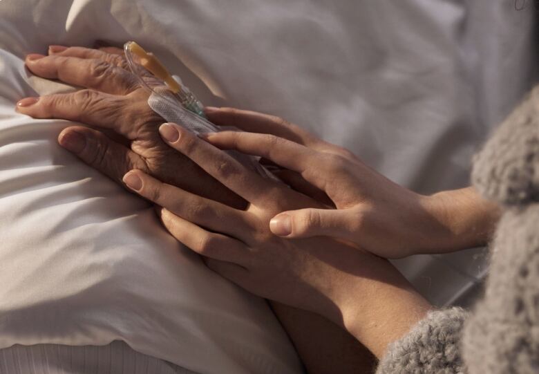 The hands of an adult hold the hand of an older person, who is hooked up to intravenous and lying in a bed.