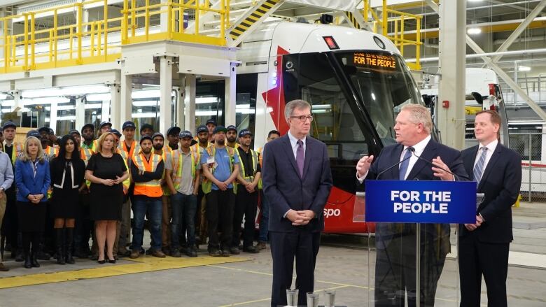 Three politicians make an announcement in a garage with a light rail train behind them.