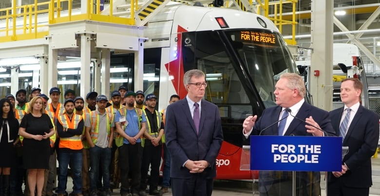 Three politicians make an announcement in a garage with a light rail train behind them.