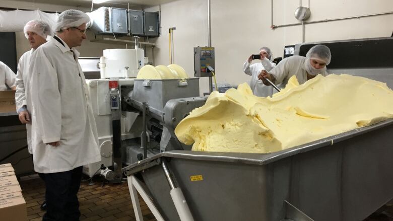 Workers clad in white jackets and hairnets shovel butter into a vat in a factory setting. 