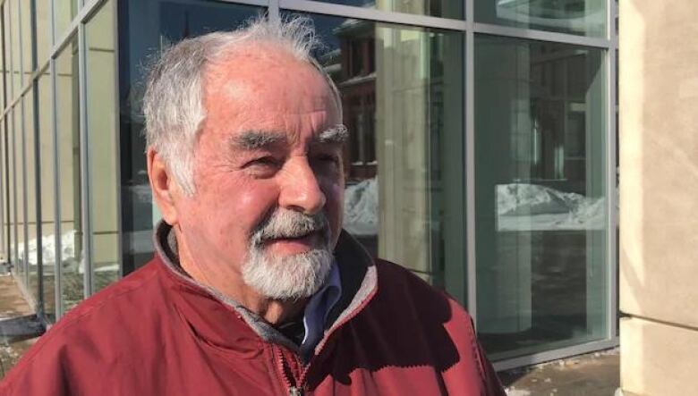 A white man in a red fleece with grey hair stands outside a building.