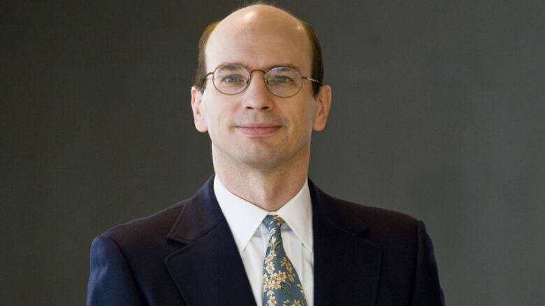 A man with round glasses wearing a suit jacket and blue and white floral tie.