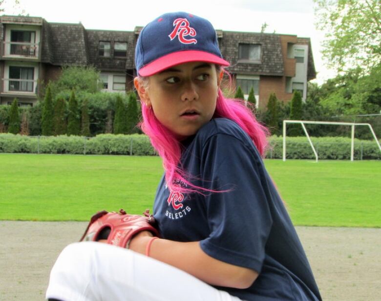 A female pitcher with bright pink hair in a pitching stance with legs raised gets ready to throw the baseball.