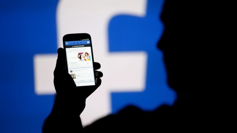 A man is silhouetted against a video screen with an Facebook logo as he poses with an Samsung S4 smartphone in this photo illustration taken in the central Bosnian town of Zenica, August 14, 2013. 