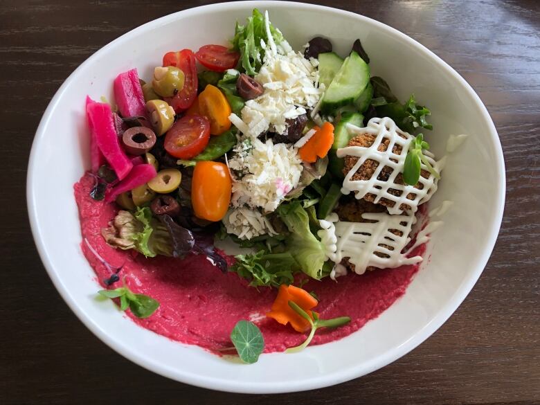 A salad featuring chery tomatoes, olives, pickled turnips, lettuce, cheese and falafel with a smear of fuschia sauce.