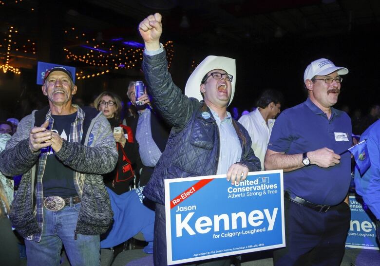 A crowd of cheering people. In the middle, is a man wearing a cowboy hat with a fist in the air. He's holding a sign that says Kenney.