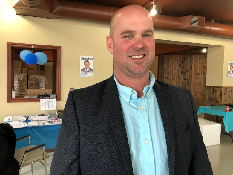 Man in sports jacket with light teal-blue shirt stands in a Progressive Conservative campaign room. 