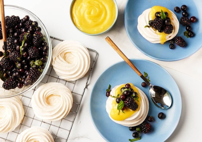 Overhead shot of mini pavlovas topped with lemon curd and fresh berries on blue dessert plates. A bowl of berries, pavlovas on a cooling rack and a bowl of the lemon curd sit next to the plates.  