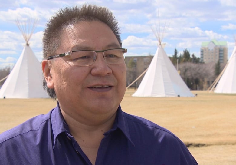 A man with glasses, wearing a purple collared shirt, stands in front of a row of teepees.