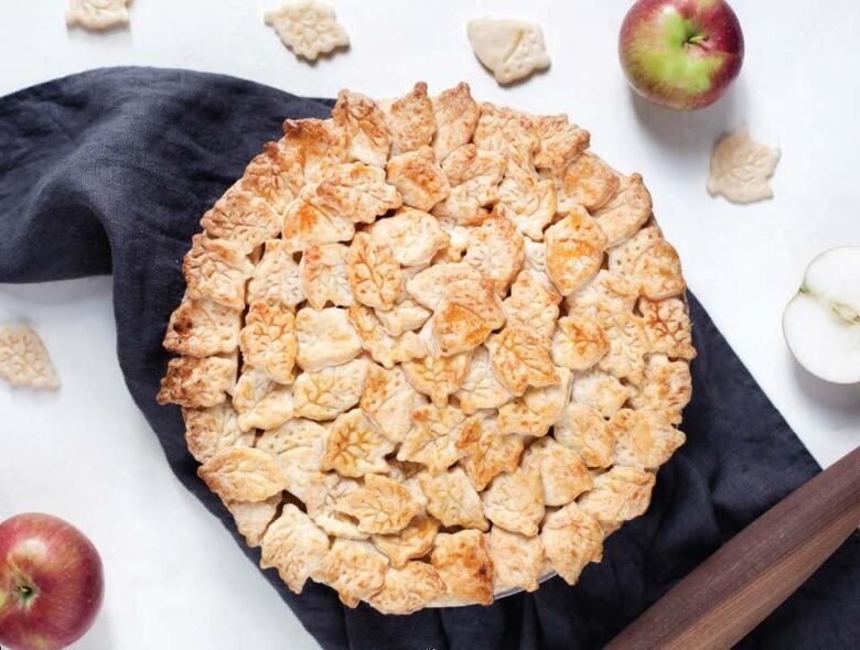 Overhead shot of an apple pie on a dark grey kitchen towel. The pie crust on top of the pie is shaped like leaves. 