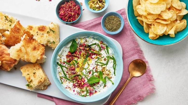 Yogurt dip garnished with pistachios, mint leaves and dried rose petals, in a blue bowl. The bowl is on a white platter on a pink napkin, on a white tabletop. Torn pieces of pita and a bowl of chips sit beside the dip. 