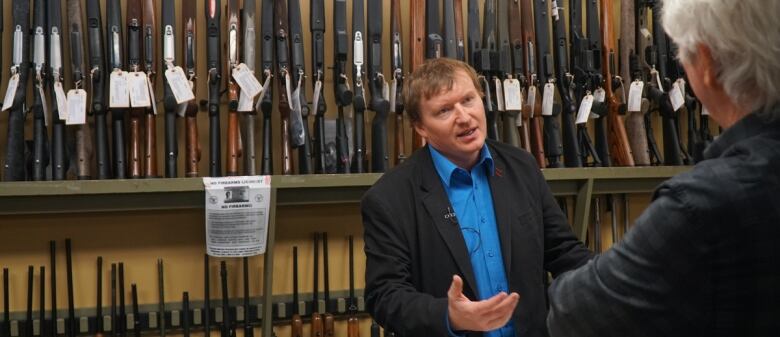 A man in a suit stands in front of a row of guns at a store.