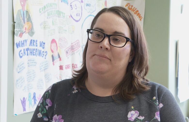 A woman wearing a flowered sweatshirt stands in front of a poster on the wall of an office.