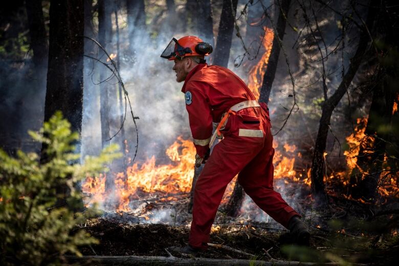 A firefighter in a red jumpsuit with soot on his face stands in smoke near a burning fire in a training exercise.