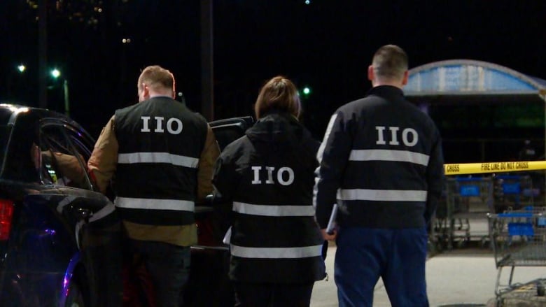 Two men stand on either side of a women and all have their back to the camera. It is dark out, but the back of their black jackets have reflector tape that spells out IIO. In front of them, is yellow police tape about waist high that says police line do not cross.