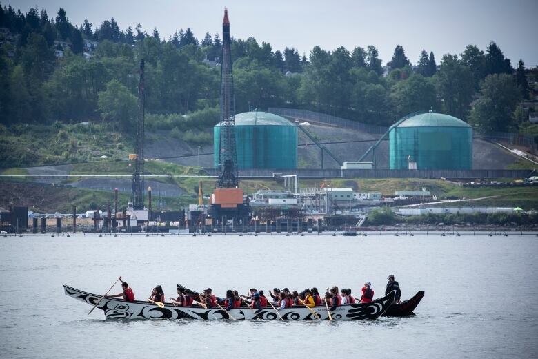 An indigenous canoe is paddled in front of oil storage tanks on the shores of Burrard Inlet.