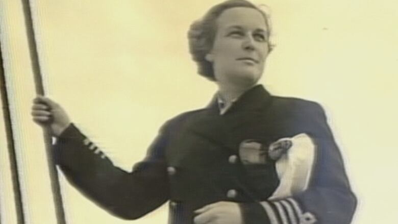 A woman in a uniform stands with one hand on the cables of a ship and the other holding her captain's hat.