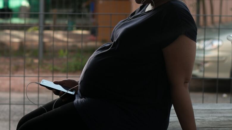 A visibly pregnant woman sits on a flat surface, holding a phone, with earbud earphones in her ears.