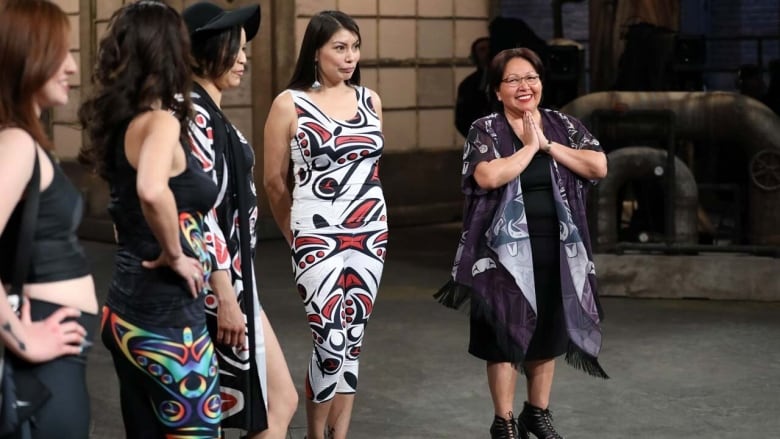 Woman smiling and holding her hands in prayer, standing beside a row of models in dresses with Gitxsan-designs on them.