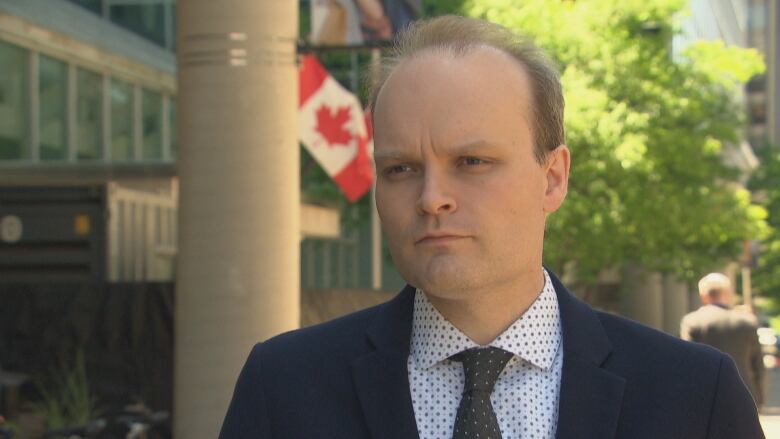 A man wearing a suit and tie stands on a leafy street in warm weather. 