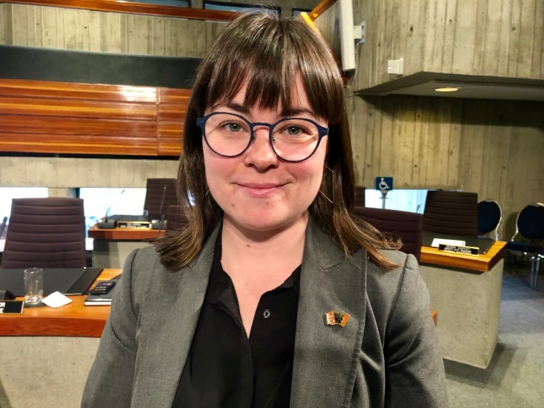 A smiling woman with short brown hair and glasses stands in a city council chambers.