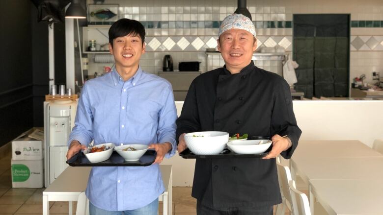 A young man and older man stand in a restaurant holding trays of bowls of food.