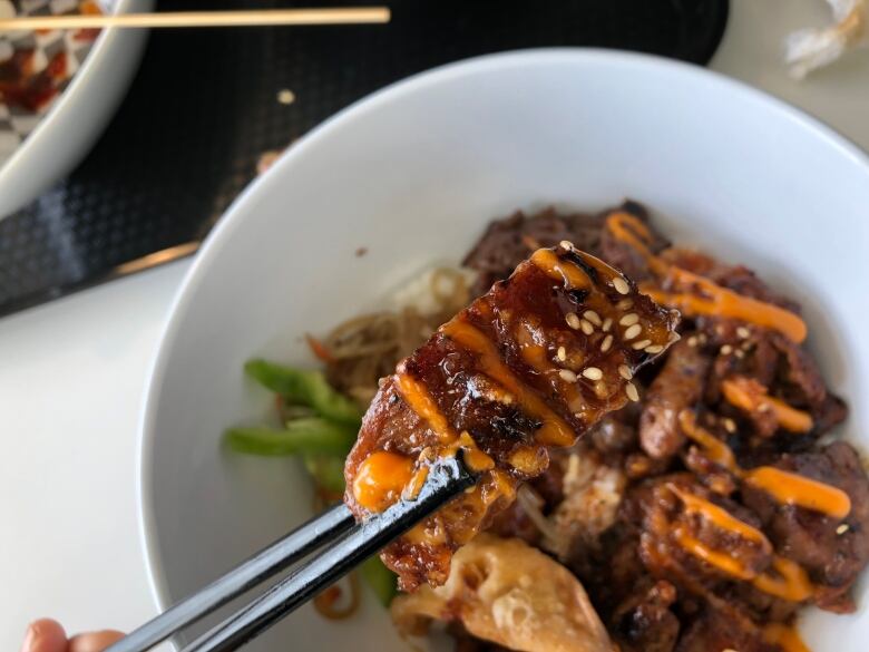 Chopsticks hold up a piece of meat drizzled with an orange sauce over a bowl filled with the same.