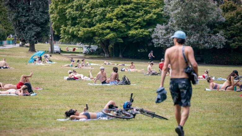 A range of people lay down on towels in a grassy area. Some of them are shirtless.