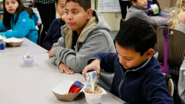 Children sitting at long tables in a school eat breakfast.