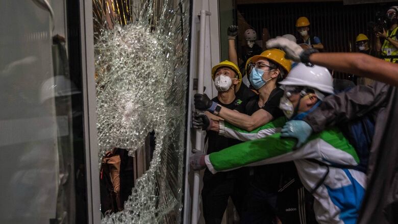 People, some wearing hard hats, smash glass on a door.