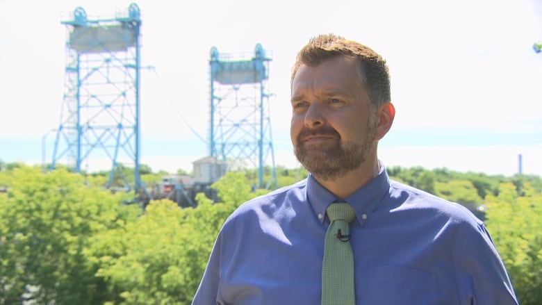 A man wearing a shirt and tie is standing outside on a sunny day, with trees in the background. 