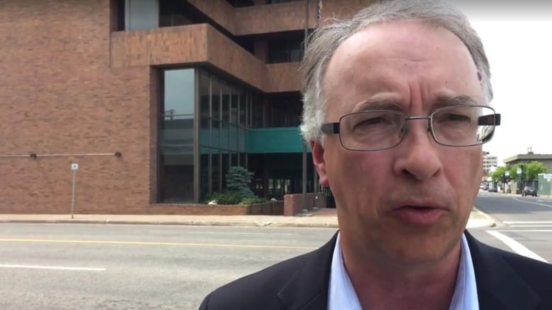 John Rustad gestures as he speaks in front of a building on a street. He's a white man with white hair and glasses.