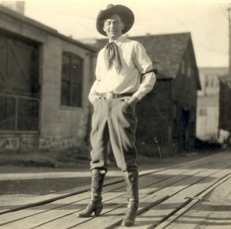 A man wearing a cowboy hat and boots stands in the street with his hands in his pockets.