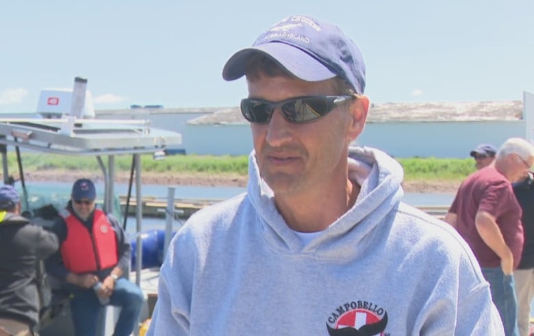 A man wearing a grey hoodie, baseball hat and sunglasses, with a boat behind him.