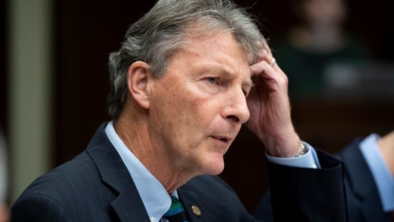 A man wearing a suit and tie scratches his head.