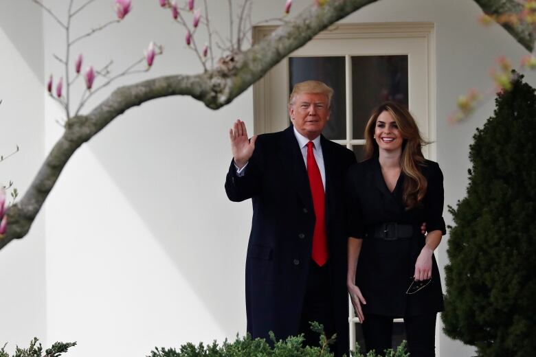 A man in a black suit with a red tie waves from the White House as he stands next to a younger woman in a black dress.