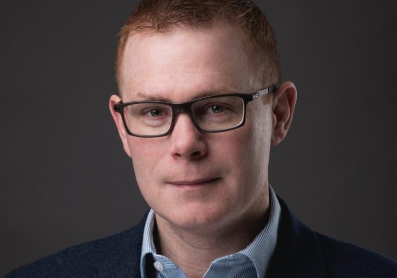 A man wearing glasses and a blue-collared shirt poses for a studio photograph.